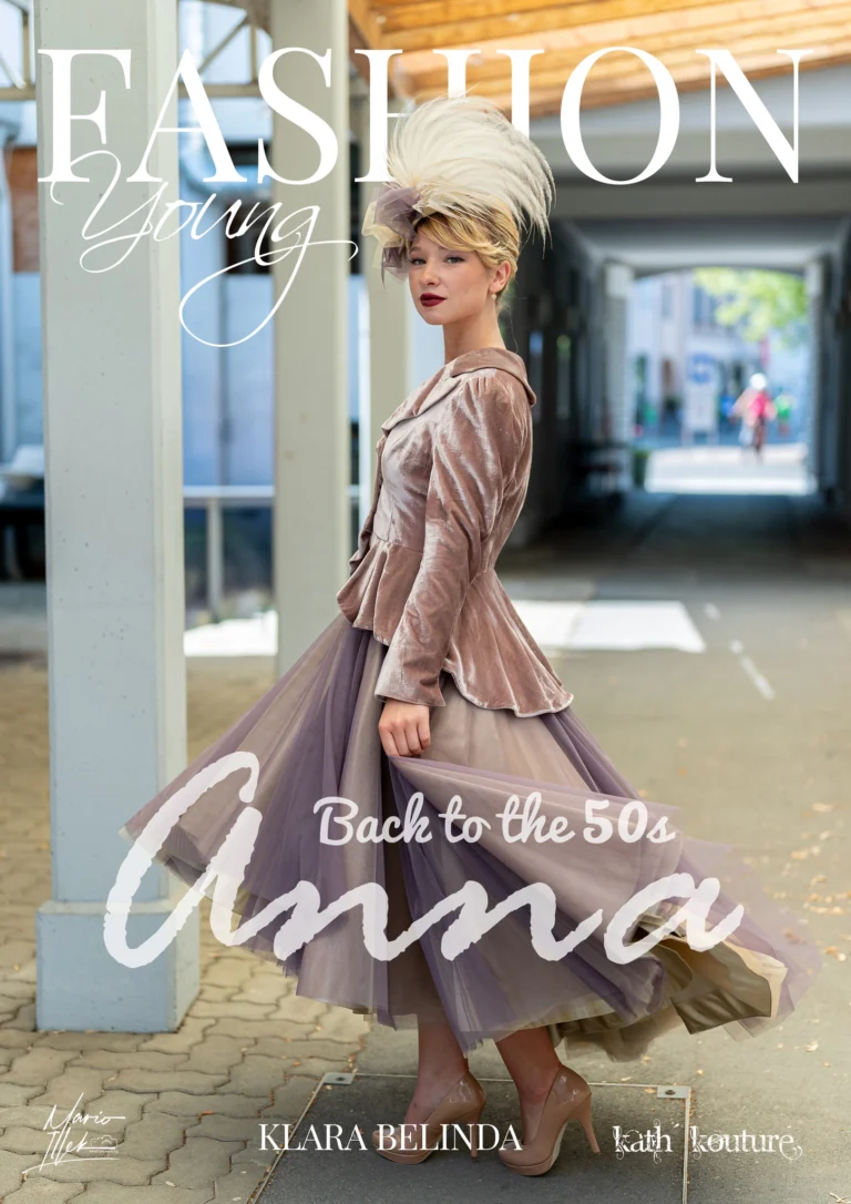 Eine Frau steht in einem überdachten Durchgang und trägt ein tailliertes, samtiges Oberteil in Rosé mit weitem Tüllrock in Violetttönen. Auf dem Kopf trägt sie einen auffälligen Kopfschmuck mit großen hellen Federn und einer Schleife. Sie blickt zur Seite, der Hintergrund zeigt eine hell beleuchtete Passage mit unscharfer Straßenszene.